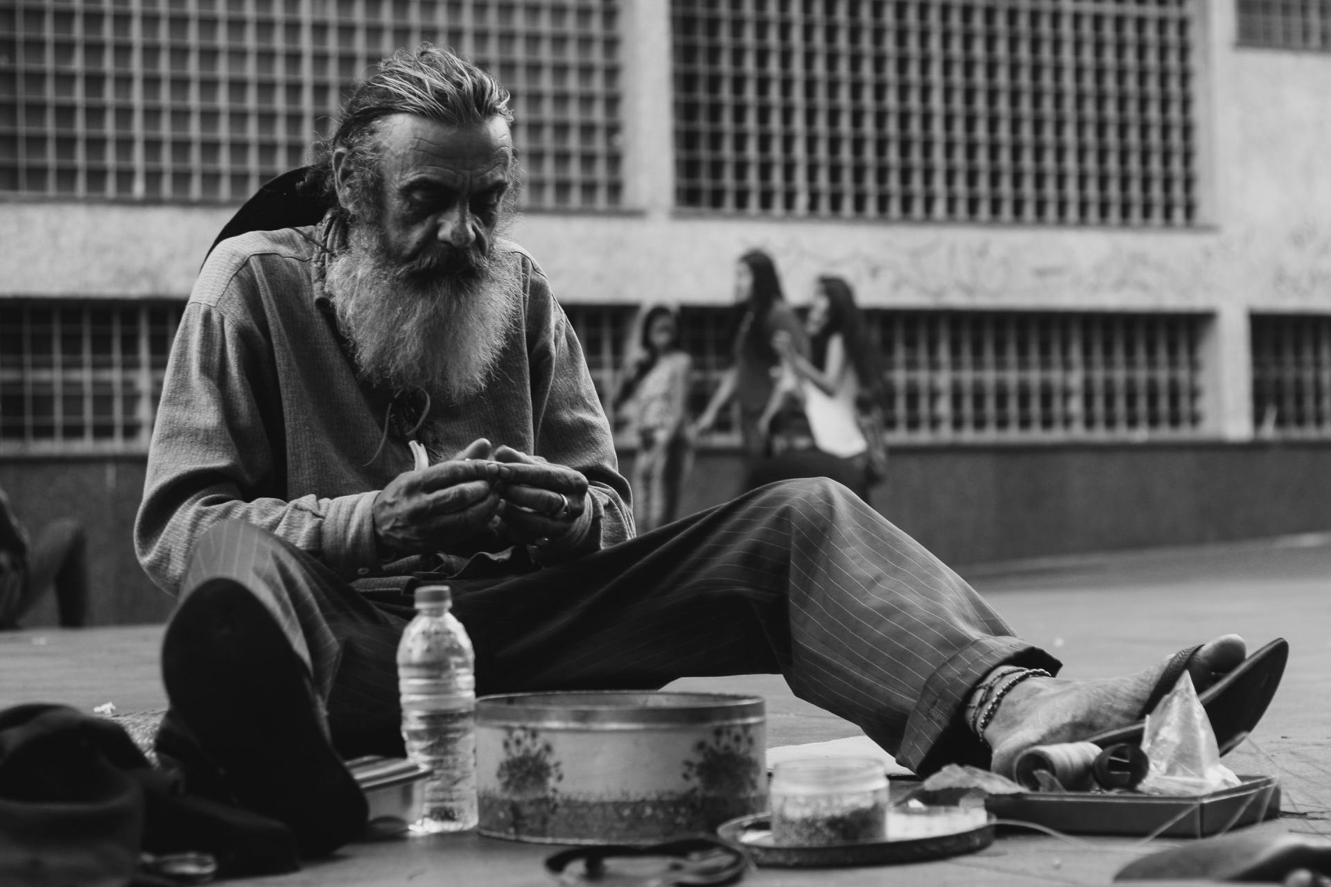 A seated man with a long beard sits beside a bottle and tray on a sidewalk, with buildings in the background.